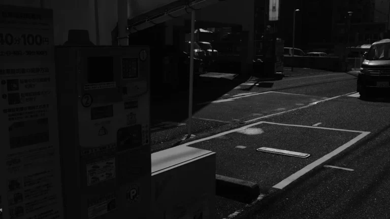 A black and white photo of a parking payment meter next to parking spaces in an urban area.