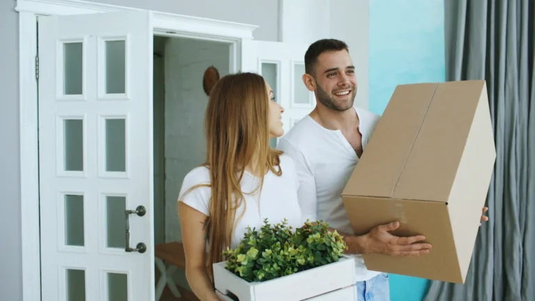 A smiling couple carrying moving boxes and a plant into their new home, representing housing security.