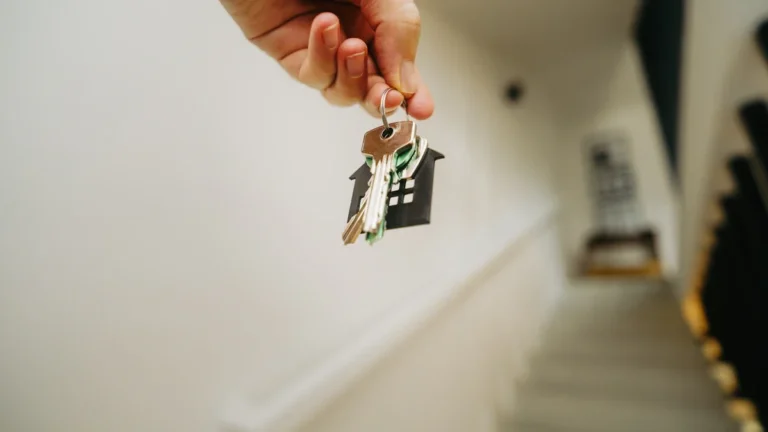 A hand holds a set of house keys with a house-shaped keychain in the hallway of a residential building.