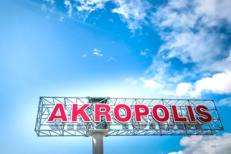 Large red letters spelling AKROPOLIS on a metal frame against a bright blue cloudy sky.