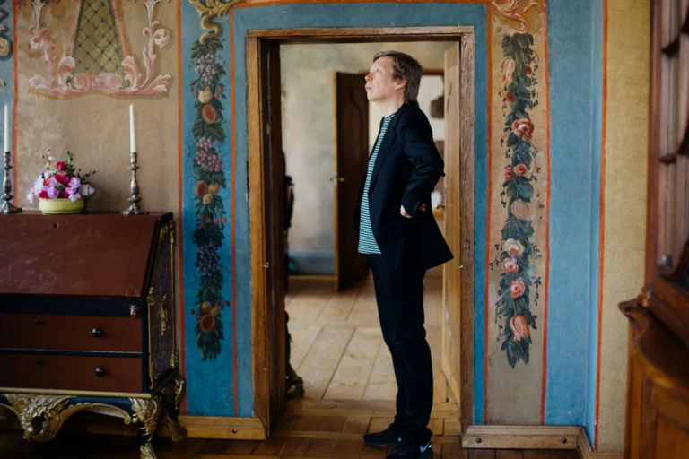 A man in a black jacket standing in a doorway of an ornate, painted historic room.