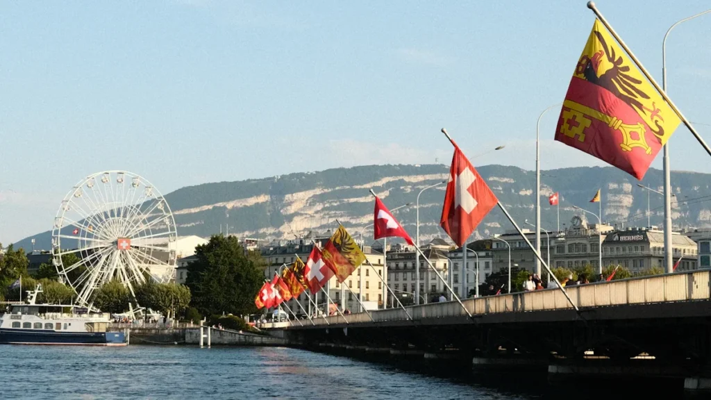 View of the Mont-Blanc Bridge in Geneva, Switzerland, featuring waving flags and a large ferris wheel.