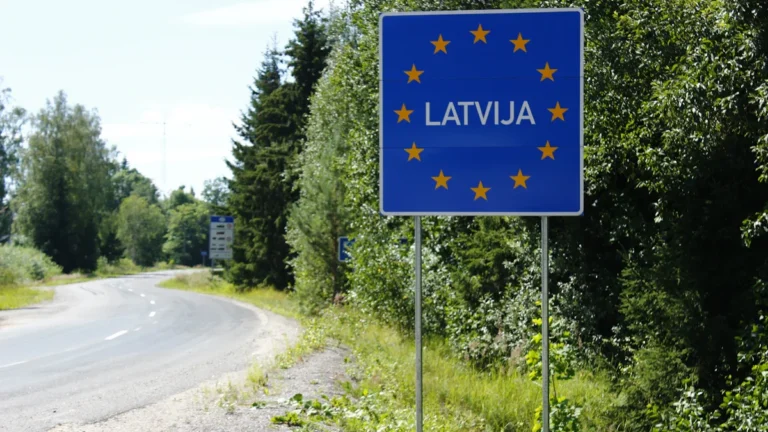 A blue European Union border sign for Latvia stands alongside a quiet road through a forest.