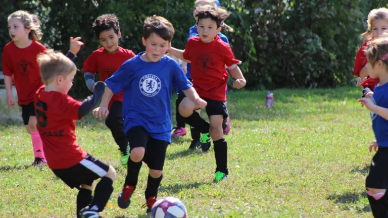 Young children playing soccer on a grass field, with one boy wearing a blue Chelsea FC jersey.