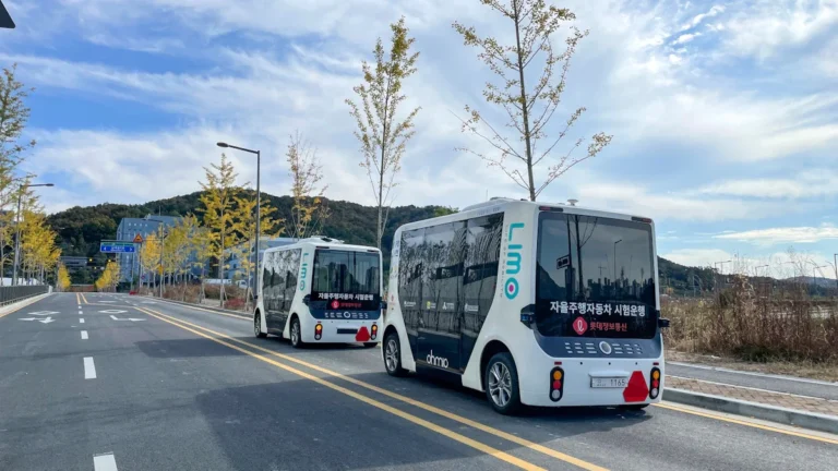 Two white autonomous shuttle buses driving in tandem on a paved road during a technical trial.