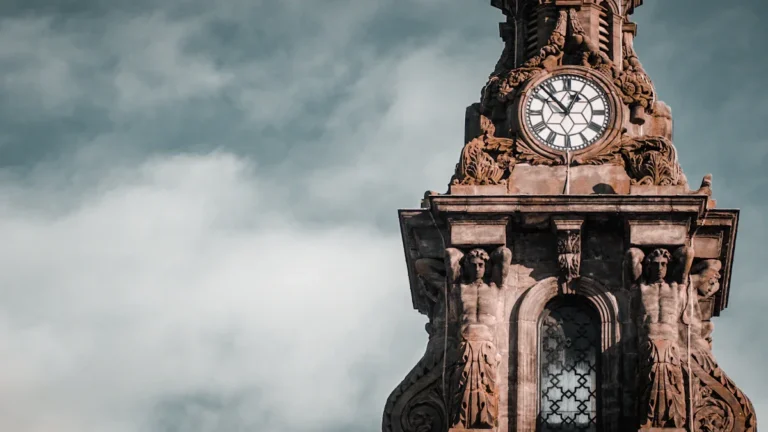 Historic stone clock tower with intricate carvings and a white face against a cloudy sky.