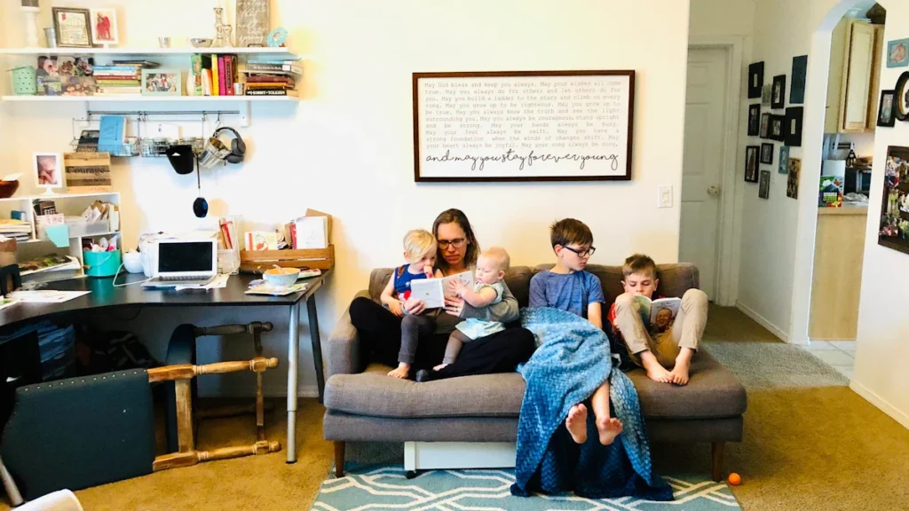A woman sits on a sofa in a cozy living room with four young children, reading and relaxing together.