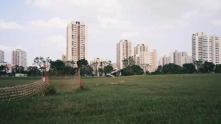 A view across a grassy field towards several high-rise residential buildings with orange construction fencing in the foreground.