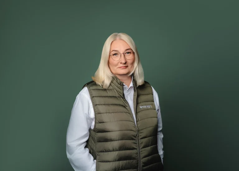 A blonde woman wearing glasses and a green branded puffer vest against a solid green background.