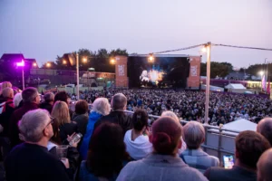 A large crowd watches an outdoor evening concert stage featuring Salt and Tar 2024 banners.