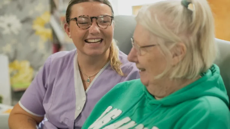 A young caregiver in purple scrubs laughs alongside an elderly woman in a green sweatshirt.