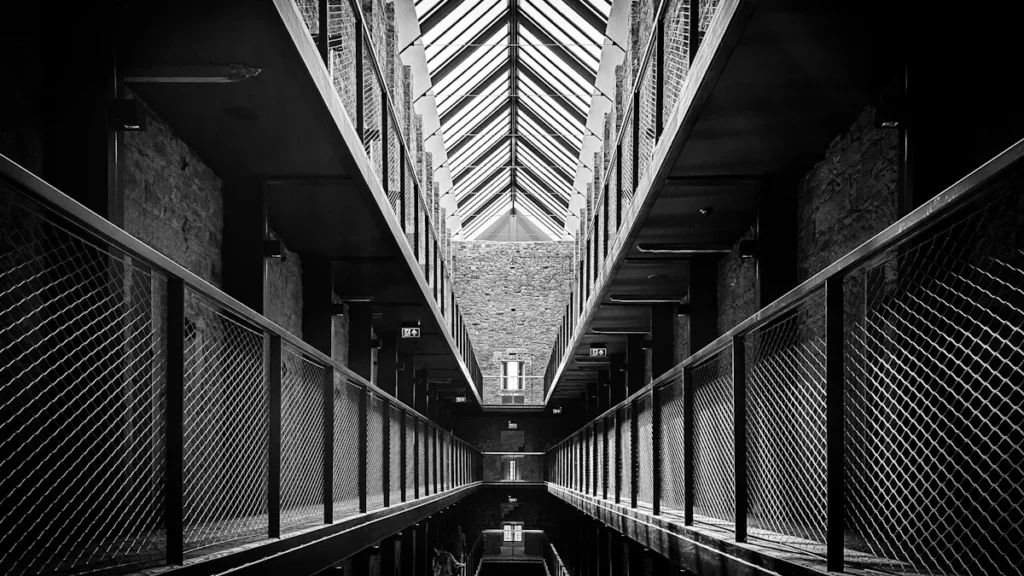 A black and white perspective view of a long, multi-level prison hallway with metal railings and skylights.