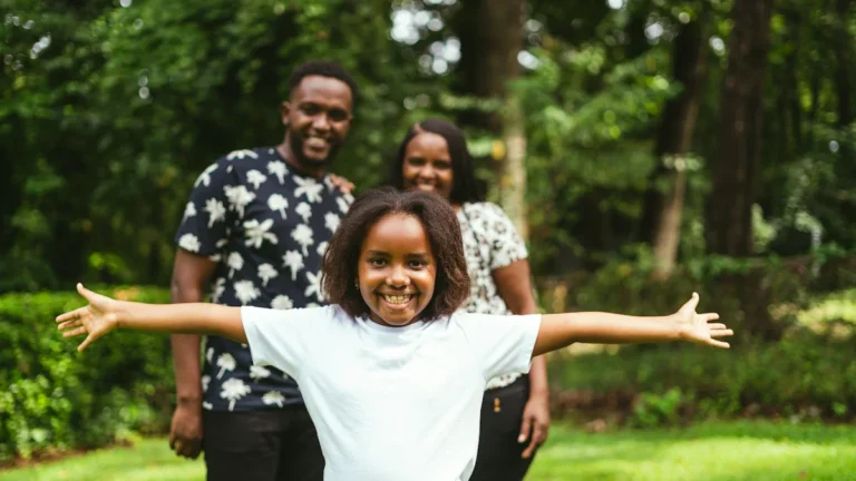 A young Black girl smiling with her arms outstretched in a park, with her foster parents standing happily in the background.