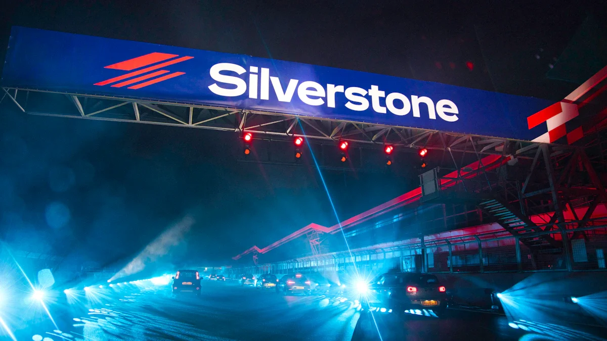 A large illuminated Silverstone sign above a race track at night with bright car headlights below.