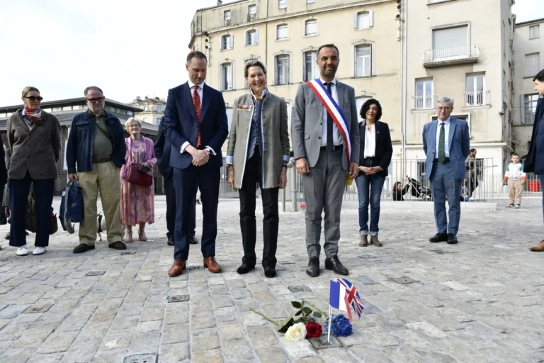Group of officials standing in a cobblestone square behind small French and British flags with flowers.