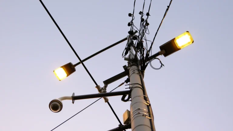 A security camera and street lights mounted on a tall pole against a pale sky.