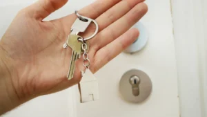 A person holding a set of house keys with a house-shaped keychain in front of a white door.