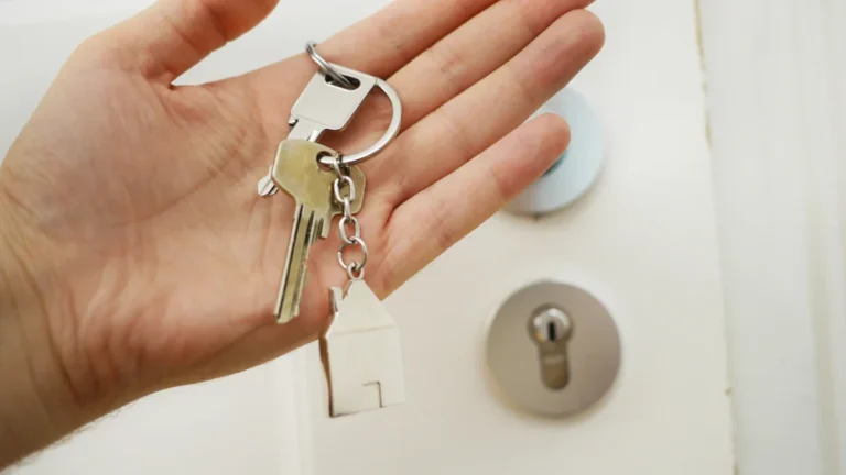 A person holding a set of house keys with a house-shaped keychain in front of a white door.