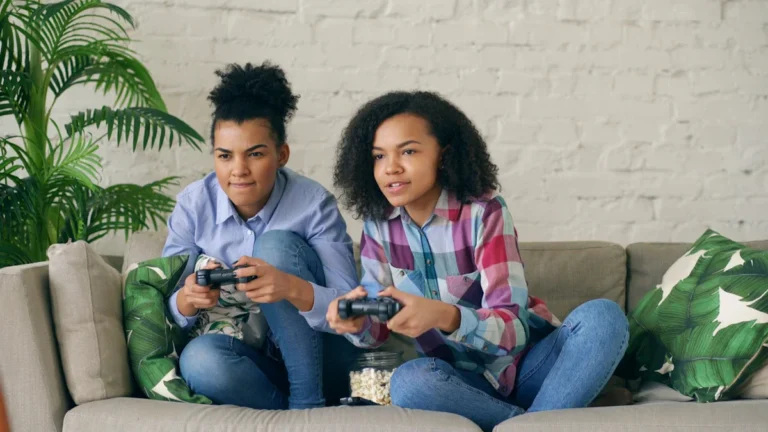 Two teenage girls sitting on a sofa playing video games together, representing the siblings and teenagers Cheshire East Council is seeking foster homes for.
