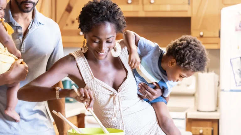 A mother smiles while stirring a bowl in a kitchen, with a young child on her hip and a father figure in the background.