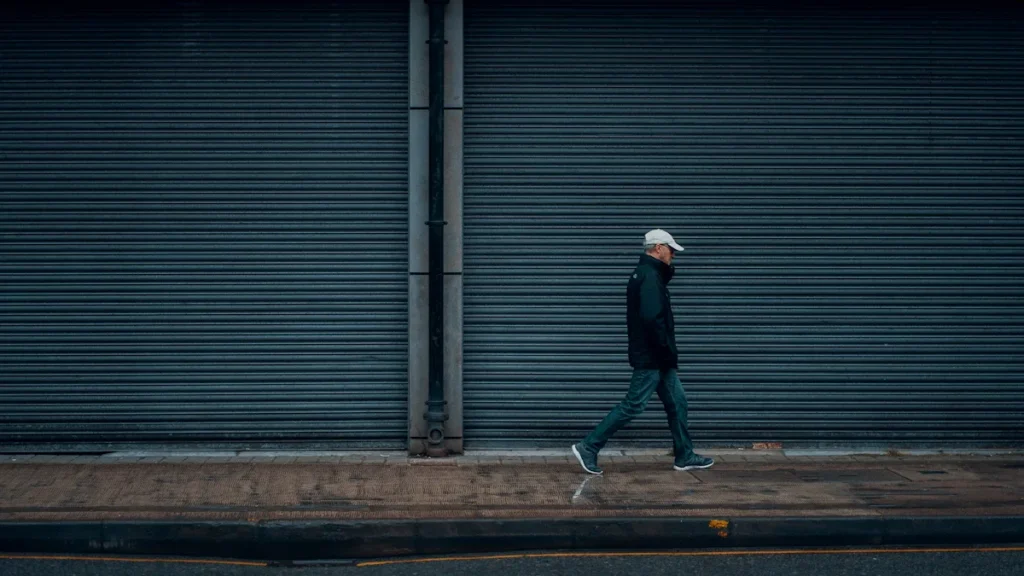 A man in a black jacket walking past closed grey metal security shutters on a city street.