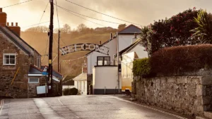 A street scene in a Cornish village featuring traditional stone houses and a road with a decorative welcome arch.