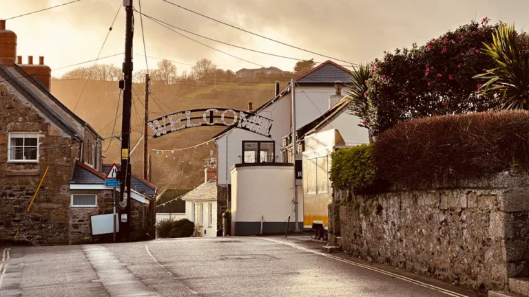 A street scene in a Cornish village featuring traditional stone houses and a road with a decorative welcome arch.