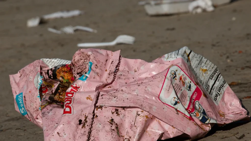 A crumpled pink snack bag and discarded food containers lying on a dirty, dusty floor.