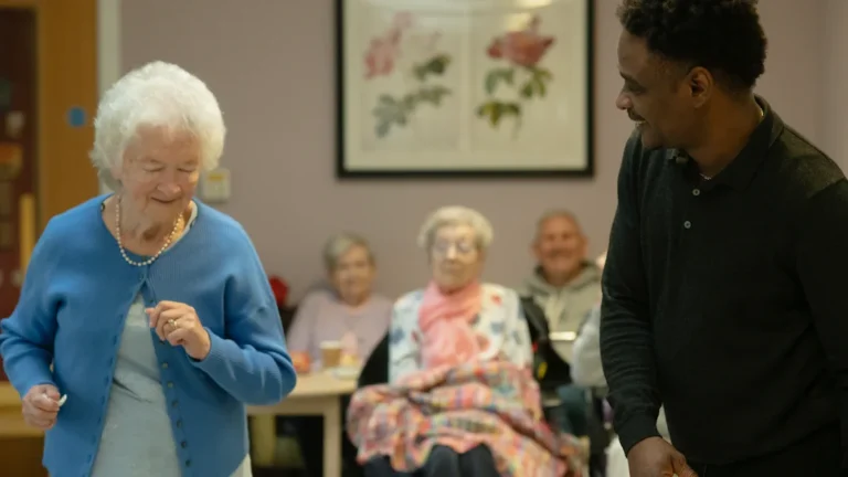 An elderly woman in a blue cardigan dances with a smiling caregiver at a UK community event.