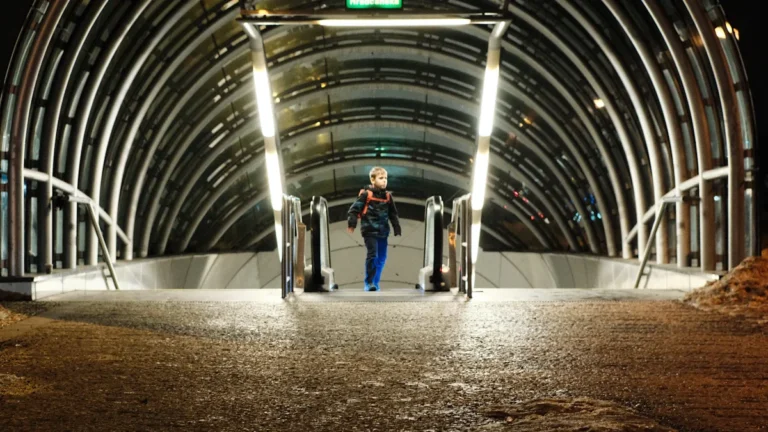 A smiling young woman in a denim jacket and yellow hijab stands in a modern, blue-lit architectural walkway.