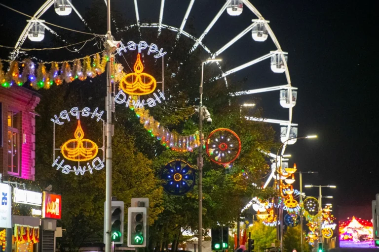 Illuminated Happy Diwali signs and a large Ferris wheel light up a festive city street.
