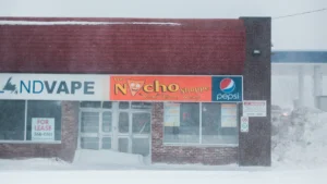 A closed-down vape shop with a 'For Lease' sign in the window during a snowstorm.
