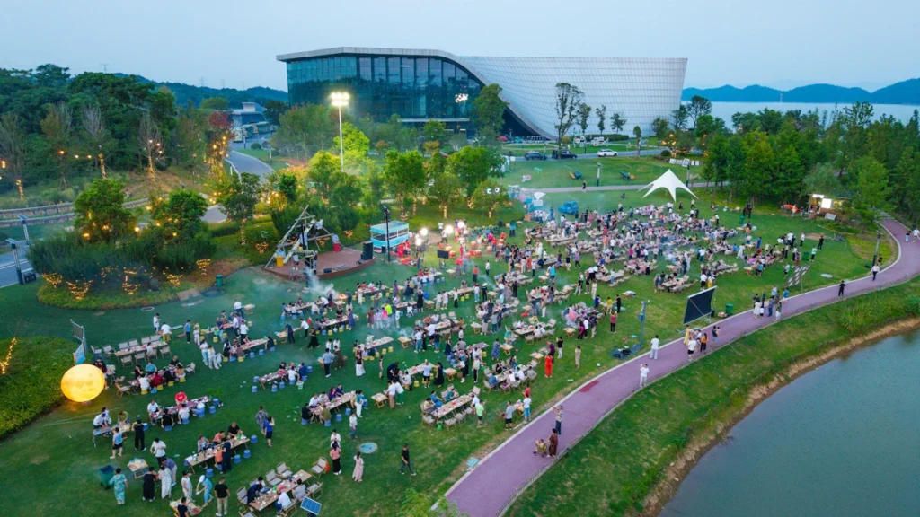 An aerial view of a massive community picnic with hundreds of attendees seated at long tables in a park beside a large modern glass building.