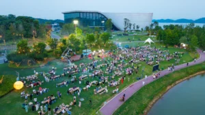An aerial view of a massive community picnic with hundreds of attendees seated at long tables in a park beside a large modern glass building.