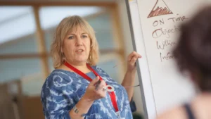 A woman in a blue patterned top teaching a class while using a white flip chart.