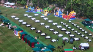 Aerial view of a large outdoor party setup in a park with rows of white tables, green marquee tents, and several colorful bouncy castles.