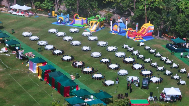 Aerial view of a large outdoor party setup in a park with rows of white tables, green marquee tents, and several colorful bouncy castles.