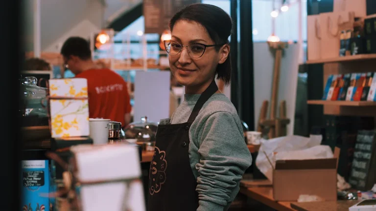 A young barista in a black apron and glasses stands behind a busy cafe counter.
