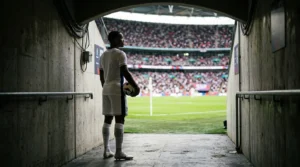 Soccer player in white uniform stands in a tunnel looking out at a packed stadium.