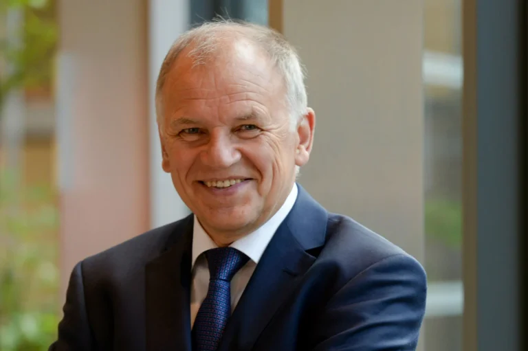 Smiling man with grey hair wearing a navy blue suit, white shirt, and patterned tie.