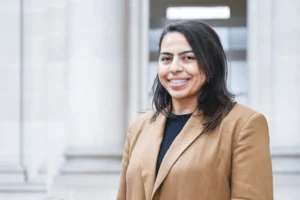 Smiling woman with dark hair wearing a tan blazer in front of blurred architectural columns.