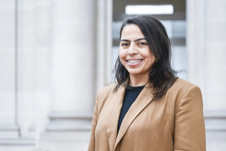 Smiling woman with dark hair wearing a tan blazer in front of blurred architectural columns.