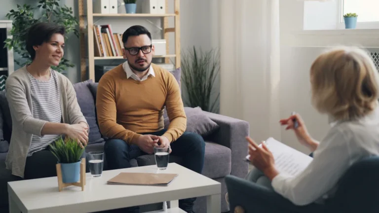 A young couple sits on a couch during a professional counseling session with a therapist in a bright office.