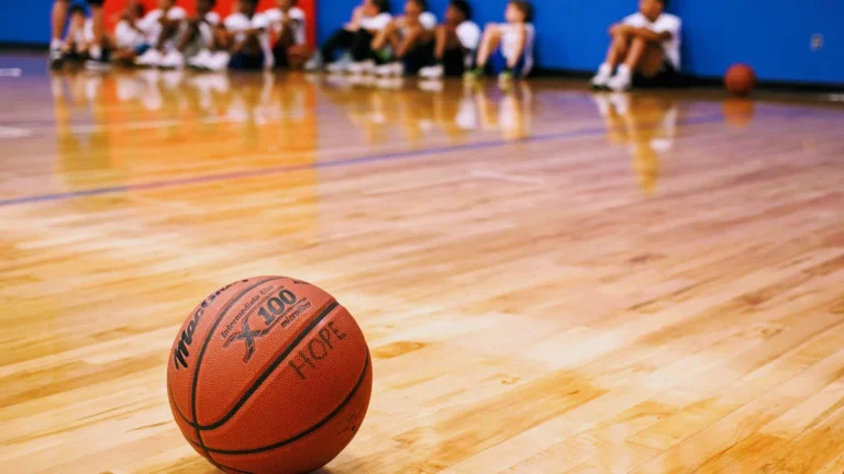 A basketball sits on a polished indoor wooden court with children sitting in the blurred background.