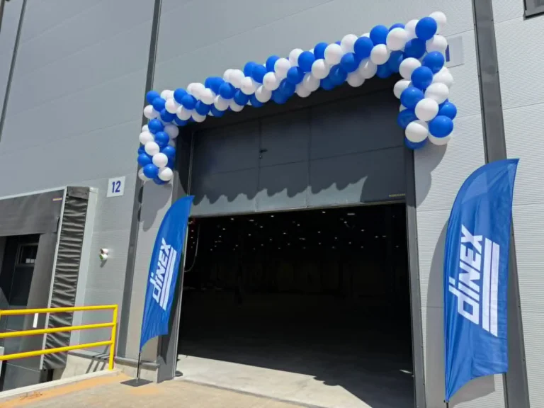 Blue and white balloon arch frames a warehouse entrance with two Dinex branded flags.