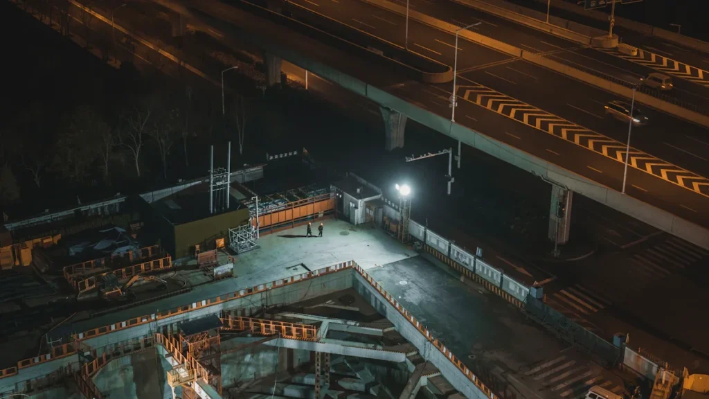 A high-angle nighttime view of an illuminated urban construction site with deep excavations and structural supports.