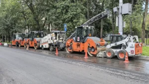 A line of heavy road resurfacing machinery, including rollers and pavers, parked on a city street in preparation for a major infrastructure project.