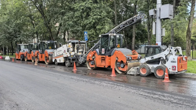 A line of heavy road resurfacing machinery, including rollers and pavers, parked on a city street in preparation for a major infrastructure project.