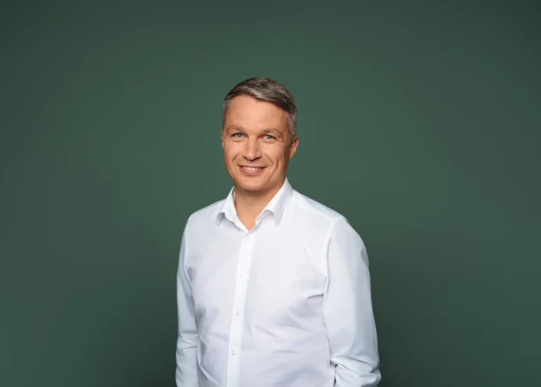 Smiling middle-aged man in a white button-down shirt against a solid dark green background.