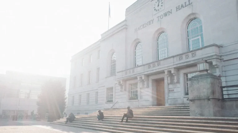 The exterior of Hackney Town Hall with 'HACKNEY TOWN HALL' visible on the facade, bathed in bright sunlight with people sitting on the steps.